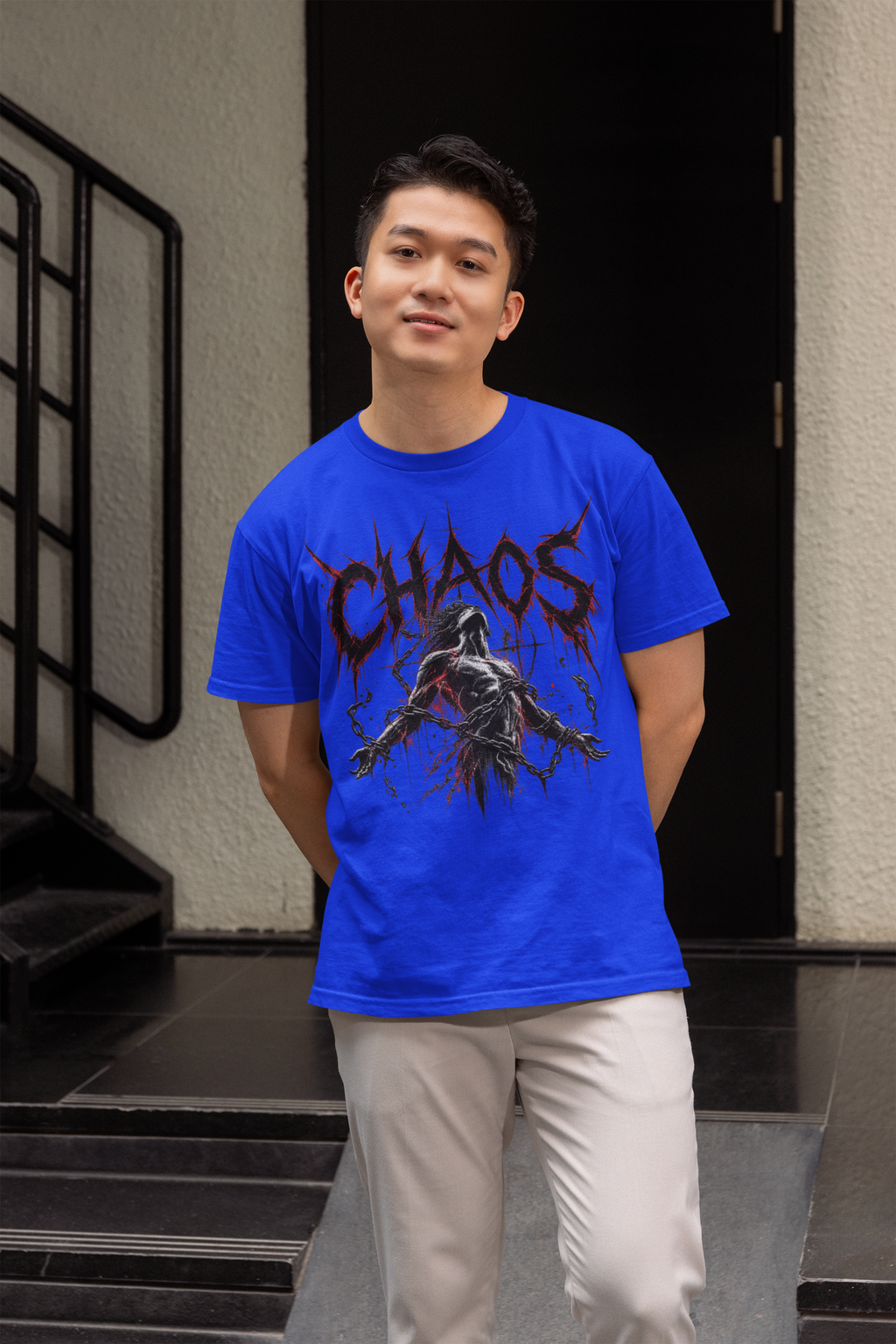 A man wearing a bright blue graphic t-shirt with a 'CHAOS' horror character print, paired with light beige trousers, standing in front of a dark doorway.