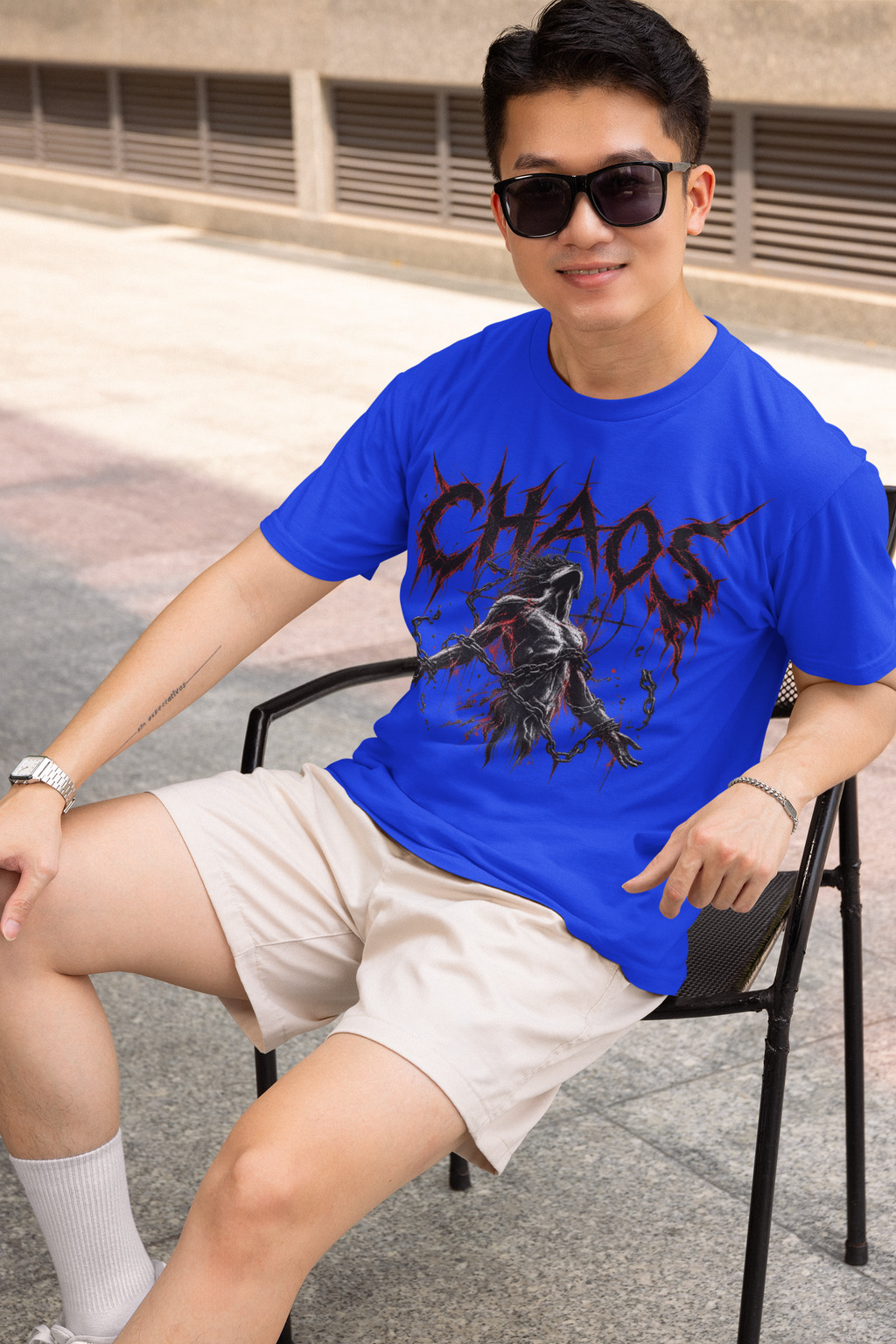 A man wearing a bright blue graphic t-shirt with the word 'CHAOS' and a chaotic character print, paired with beige shorts and sunglasses, sitting on a black chair outdoors.