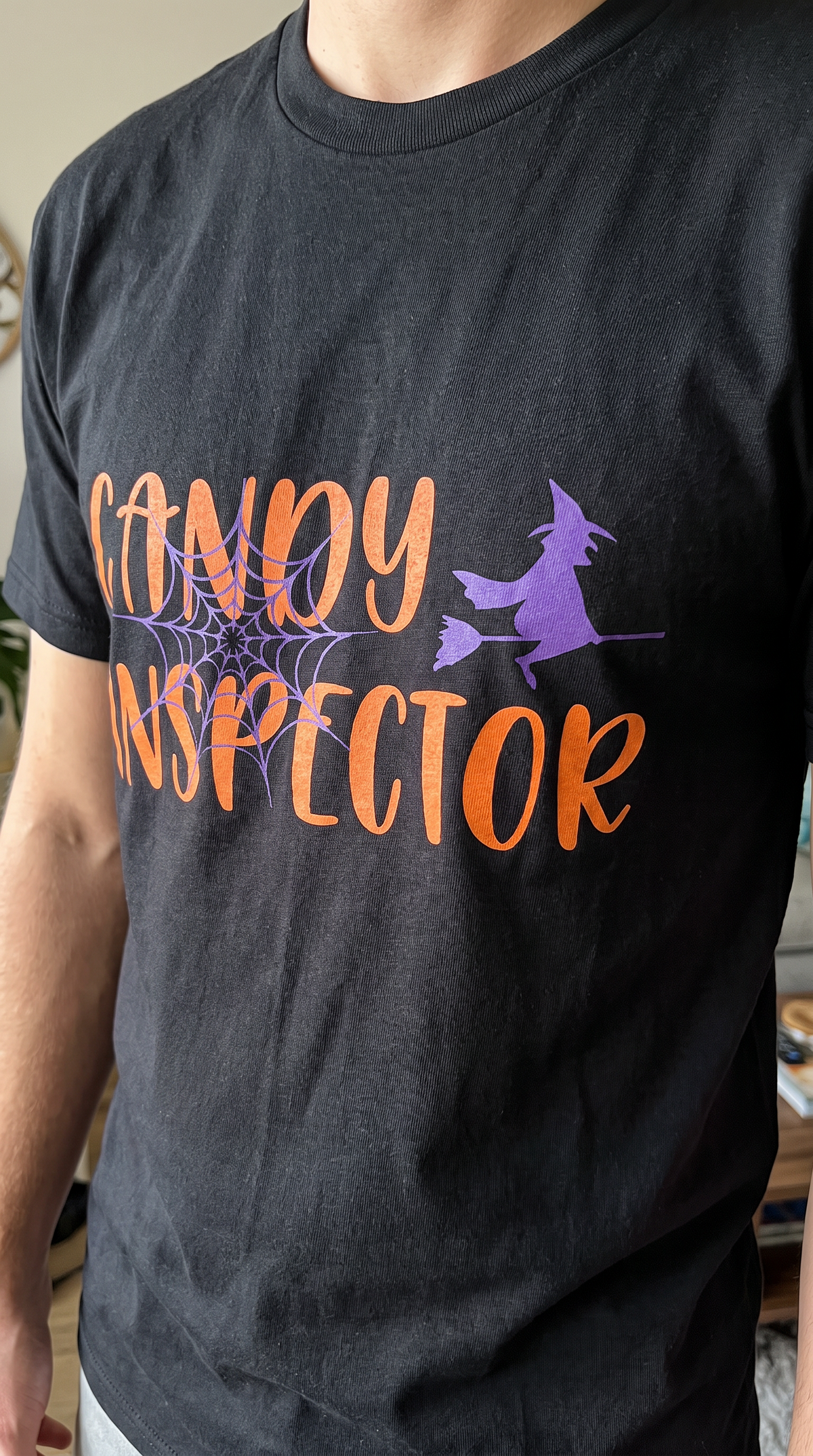 Person wearing the black Candy Inspector Halloween tee with orange CANDY INSPECTOR lettering, spider web, and a purple witch
