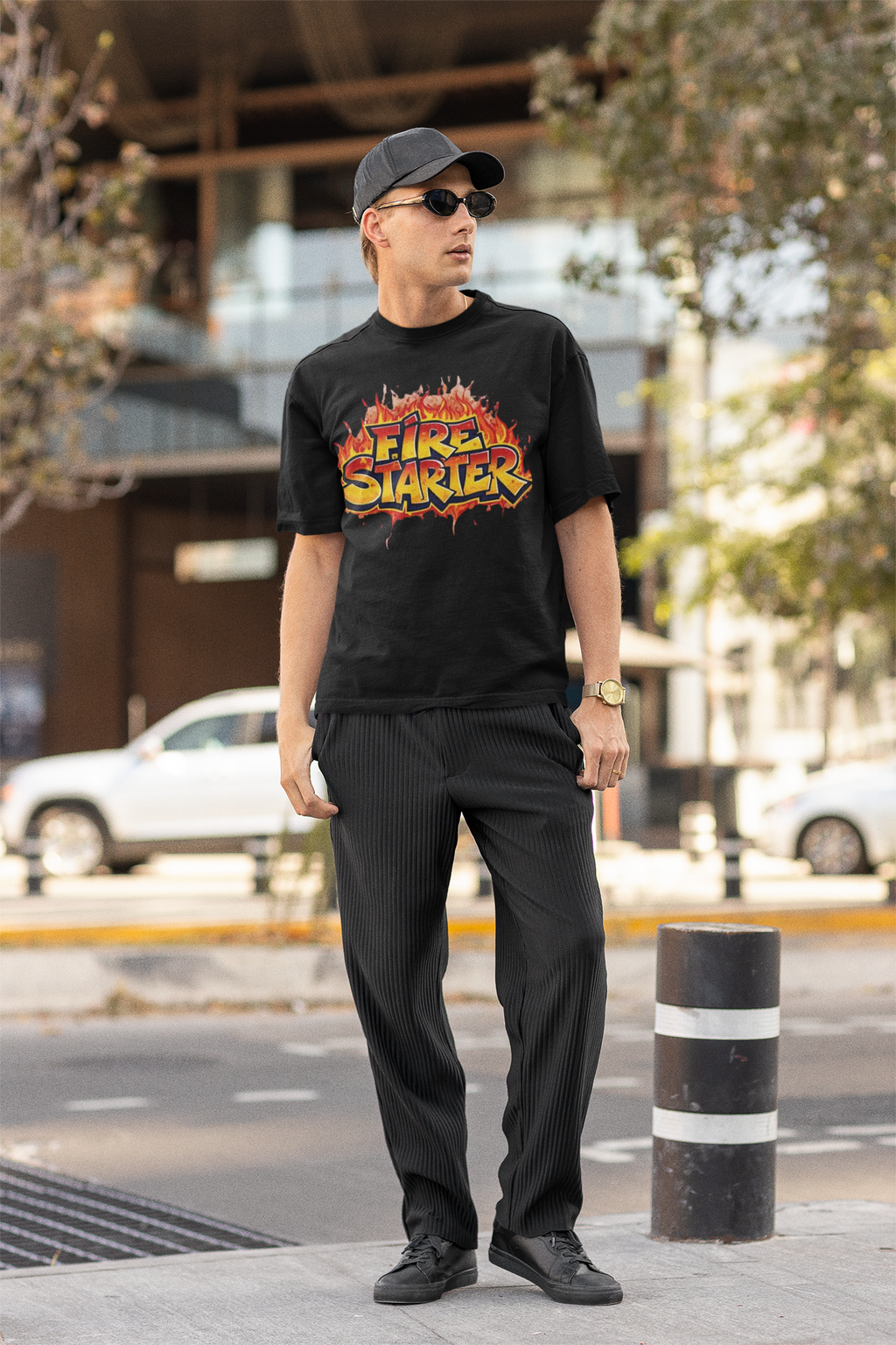 A man stands on a city sidewalk wearing a black graphic T-shirt with a large flame-style 'Revel Starter' print, black pinstriped trousers, black shoes, a dark baseball cap, and sunglasses. The background shows a blurred urban street scene with buildings, trees, parked cars, and a bollard.