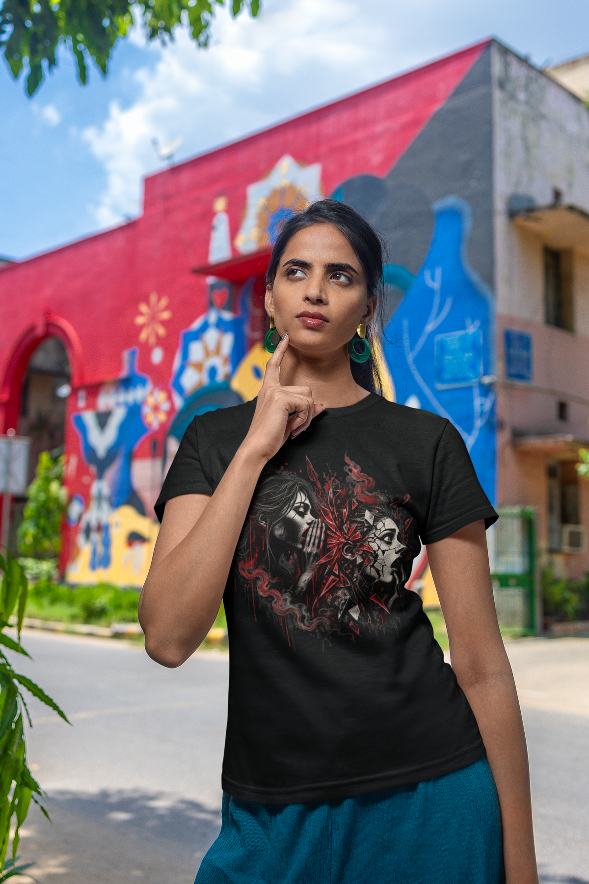 A woman wearing a black graphic t-shirt with a red and white skull design, posing outdoors in front of a vibrant red and blue painted building.