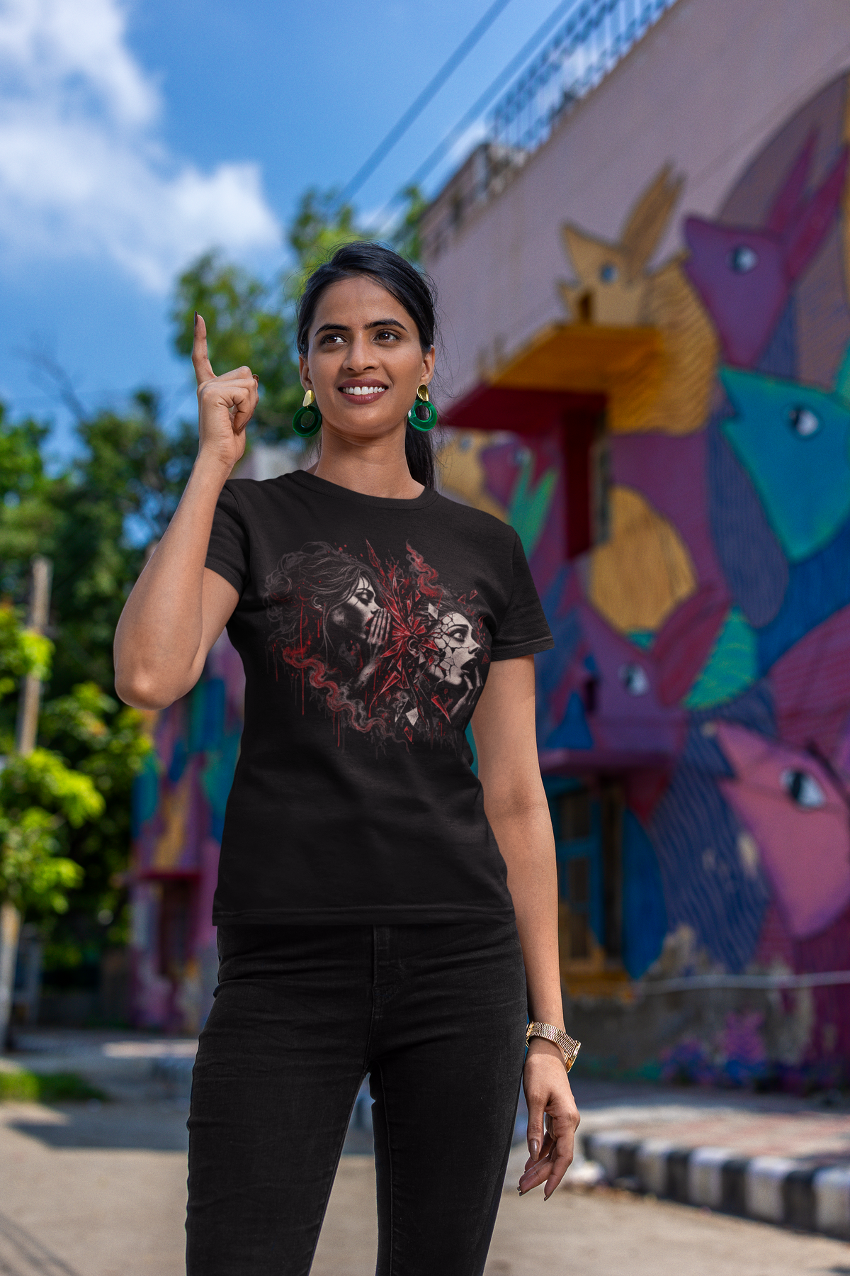 A woman wearing a black graphic t-shirt with a skull and face print, posing outdoors in front of a colorful graffiti wall.