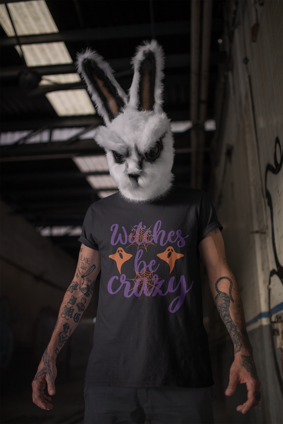 A person wearing a white bunny mask and arm tattoos stands in a dim industrial interior, wearing a black graphic T-shirt with purple script text reading 'Witches be crazy' and orange ghost illustrations.